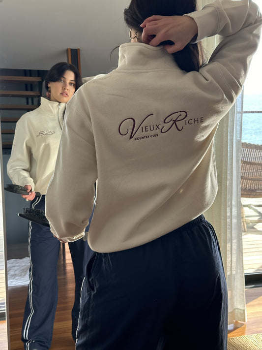 Back view of a woman wearing Vieux Riche Heritage Club half-zip sweatshirt in ivory, looking at her reflection in the mirror.