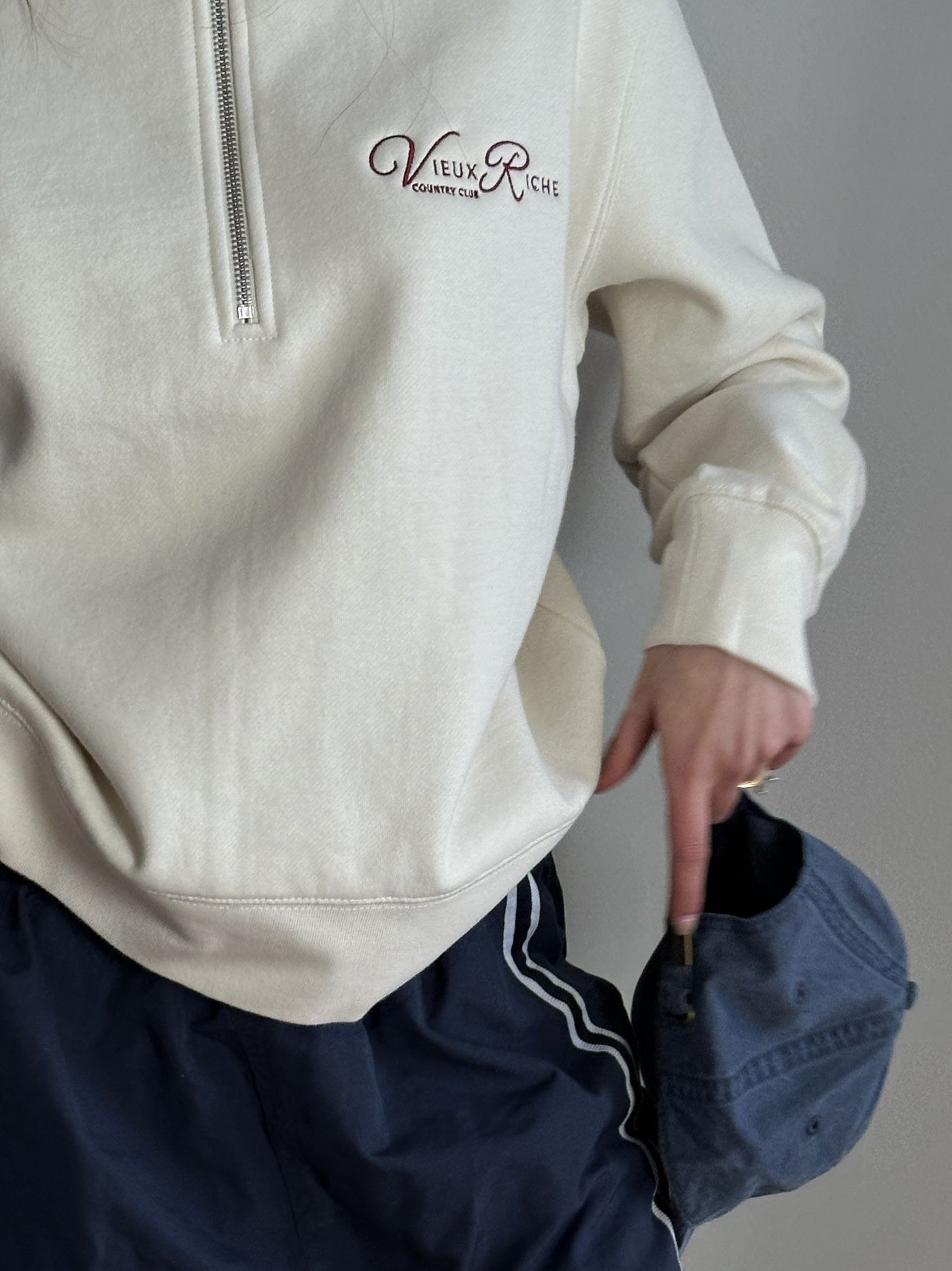 Close-up of Vieux Riche Heritage Club half-zip sweatshirt in ivory, showing the front logo and a navy cap.