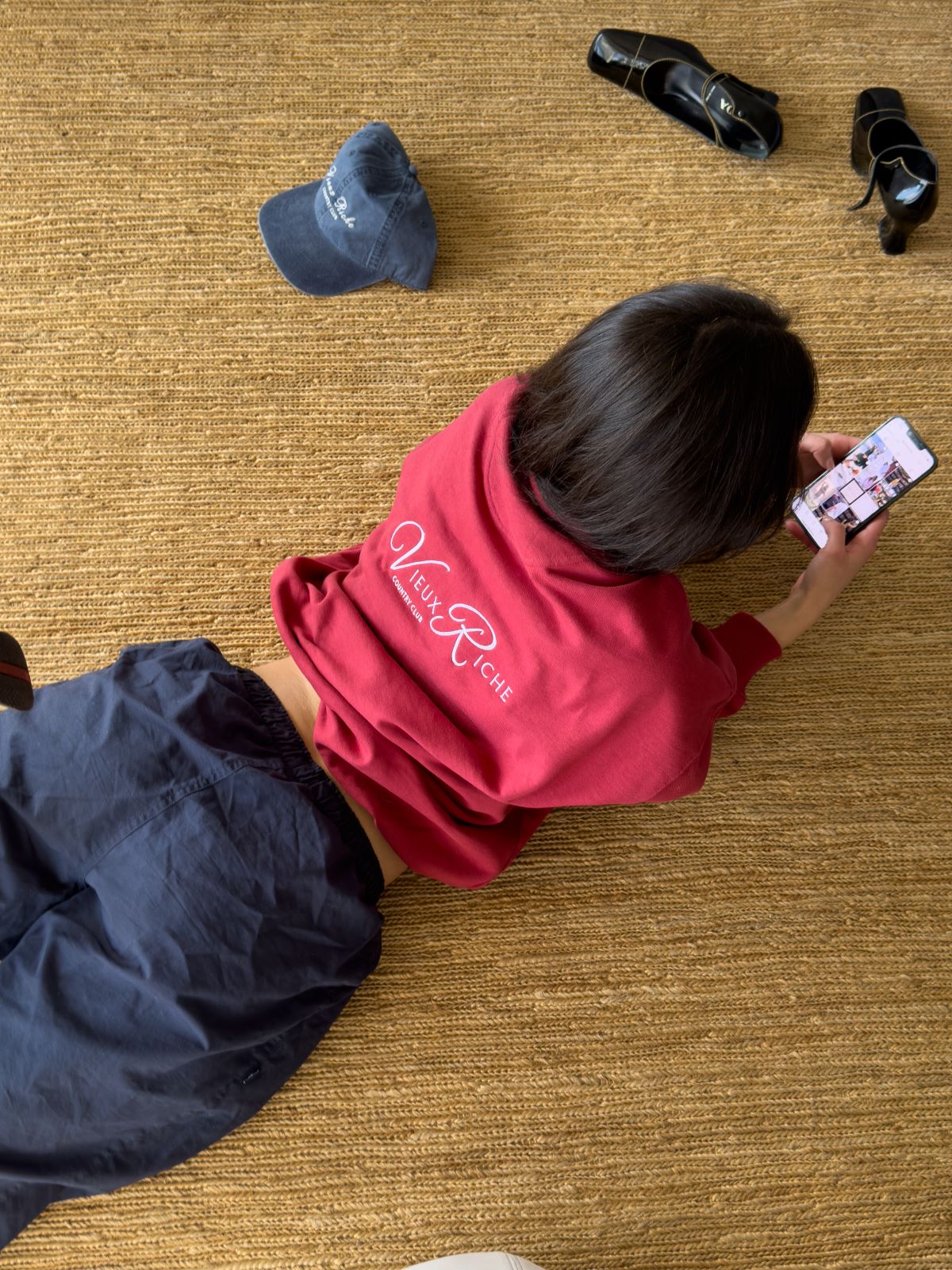 Back view of a woman wearing Vieux Riche Heritage Club half-zip sweatshirt in maroon, lying on the floor with a phone.