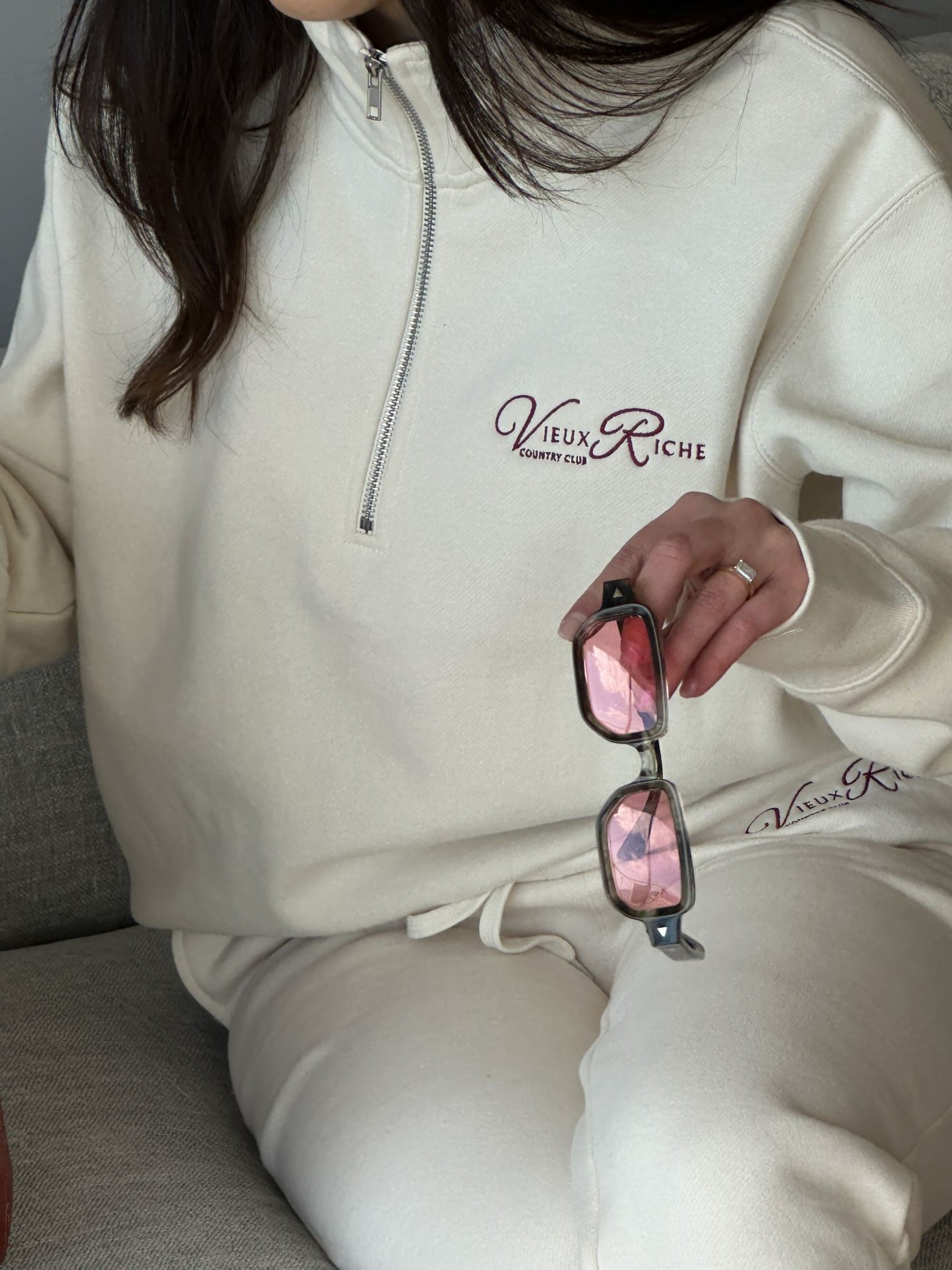 Close-up of Vieux Riche Heritage Club track pants in ivory with logo visible, worn with a matching half-zip sweatshirt and holding sunglasses.