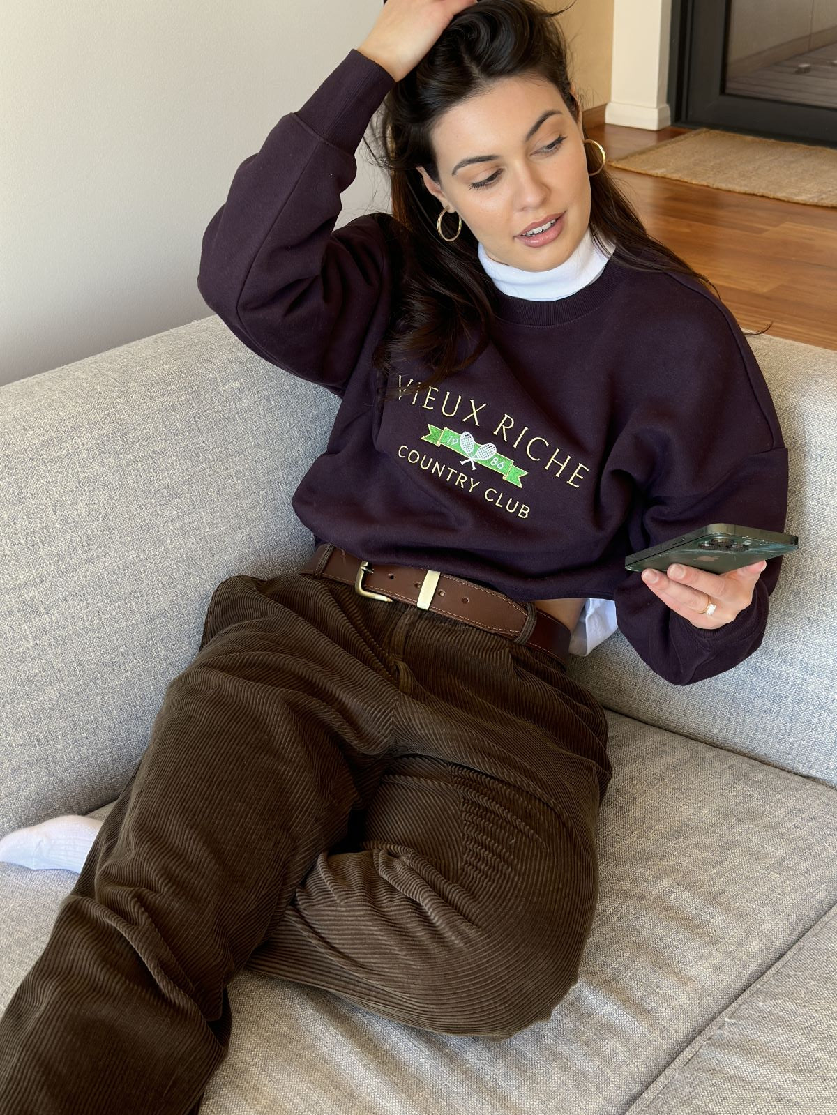 Woman in Vieux Riche Vintage Country Club burgundy sweatshirt with brown corduroy trousers, sitting on couch.